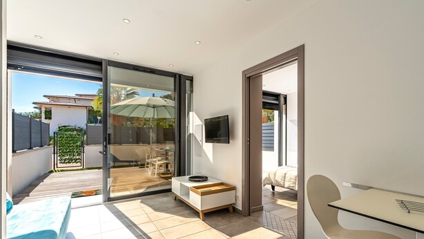 A modern living room with a glass door leading to a patio, a small white table with a tray, a beige chair, and a TV mounted on the wall.