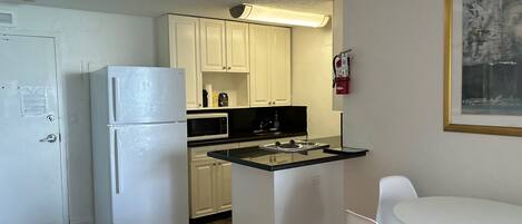 Una cocina compacta con armarios blancos, un refrigerador, un microondas y una estufa a gas. Hay un pequeño comedor con una mesa y sillas blancas.