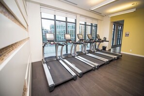 Une salle de sport où les tapis de course sont alignés le long d’un mur doté de grandes fenêtres, ce qui permet à la lumière naturelle d’entrer et offre une vue sur la ville.