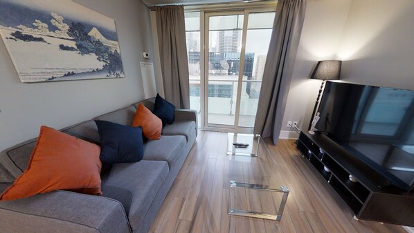 Un salon moderne avec un canapé gris, des coussins orange et bleus, une table basse en verre, un téléviseur à écran plat et une grande fenêtre donnant sur la ville.