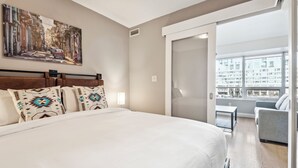 A modern hotel room with a large bed, a cityscape painting on the wall, and a view of the city through the window. - Charming 1-BR Condo in DR Toronto (Toronto)