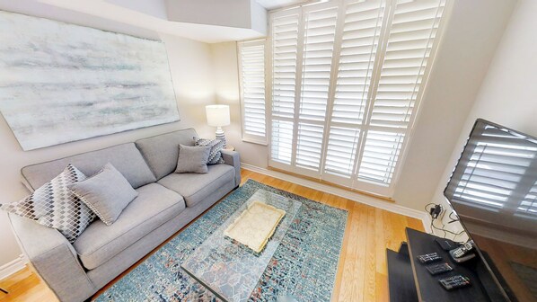A living room with a grey sofa, patterned cushions, a glass coffee table, a blue and orange rug, a TV, and a large abstract painting on the wall.