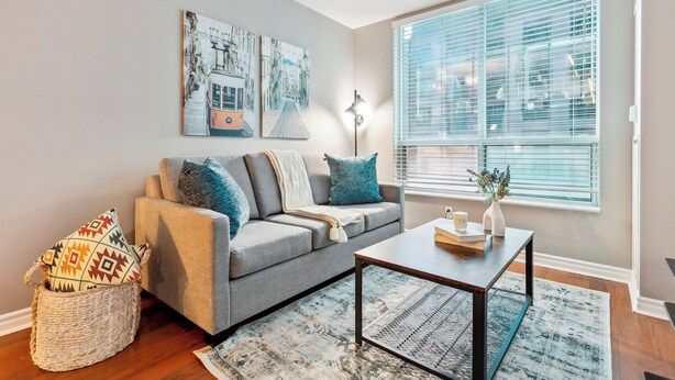 A living room with a grey sofa, patterned cushions, a woven basket, a coffee table, and a large window with blinds. - Centrally Located 1-BR Condo in DT Toronto (Toronto)