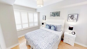 A bedroom with a large bed, white walls, and a window with blinds. There are two bedside tables with lamps and a small clock. - Centrally Located 1-BR Condo in DT Toronto (Toronto)