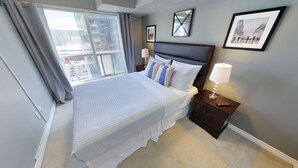 A hotel room with a large bed, a nightstand with a lamp, a framed picture on the wall, and a window with curtains. - Centrally Located 2-BR Condo in DT Toronto (Toronto)