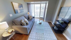 A modern living room with a grey sofa, a coffee table, a TV on a black stand, and a large window with a view of buildings. - Centrally Located 2-BR Condo in DT Toronto (Toronto)