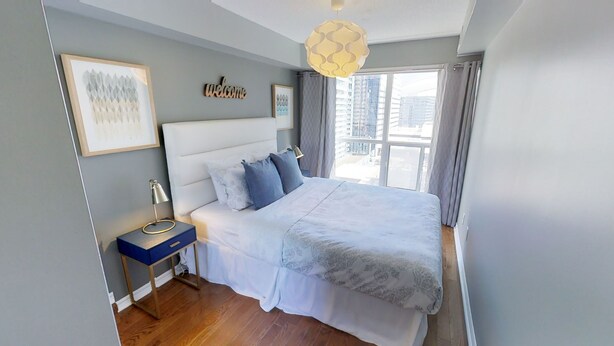 A modern bedroom with a large bed, a nightstand, a wall-mounted lamp, and a city view through the window. - Centrally Located 2-BR Condo in DT Toronto (Toronto)