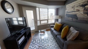 A modern living room with a flat-screen TV, a sofa with yellow cushions, a patterned rug, and a large window offering a city view. - Charming 2-BR Condo in DR Toronto (Toronto)
