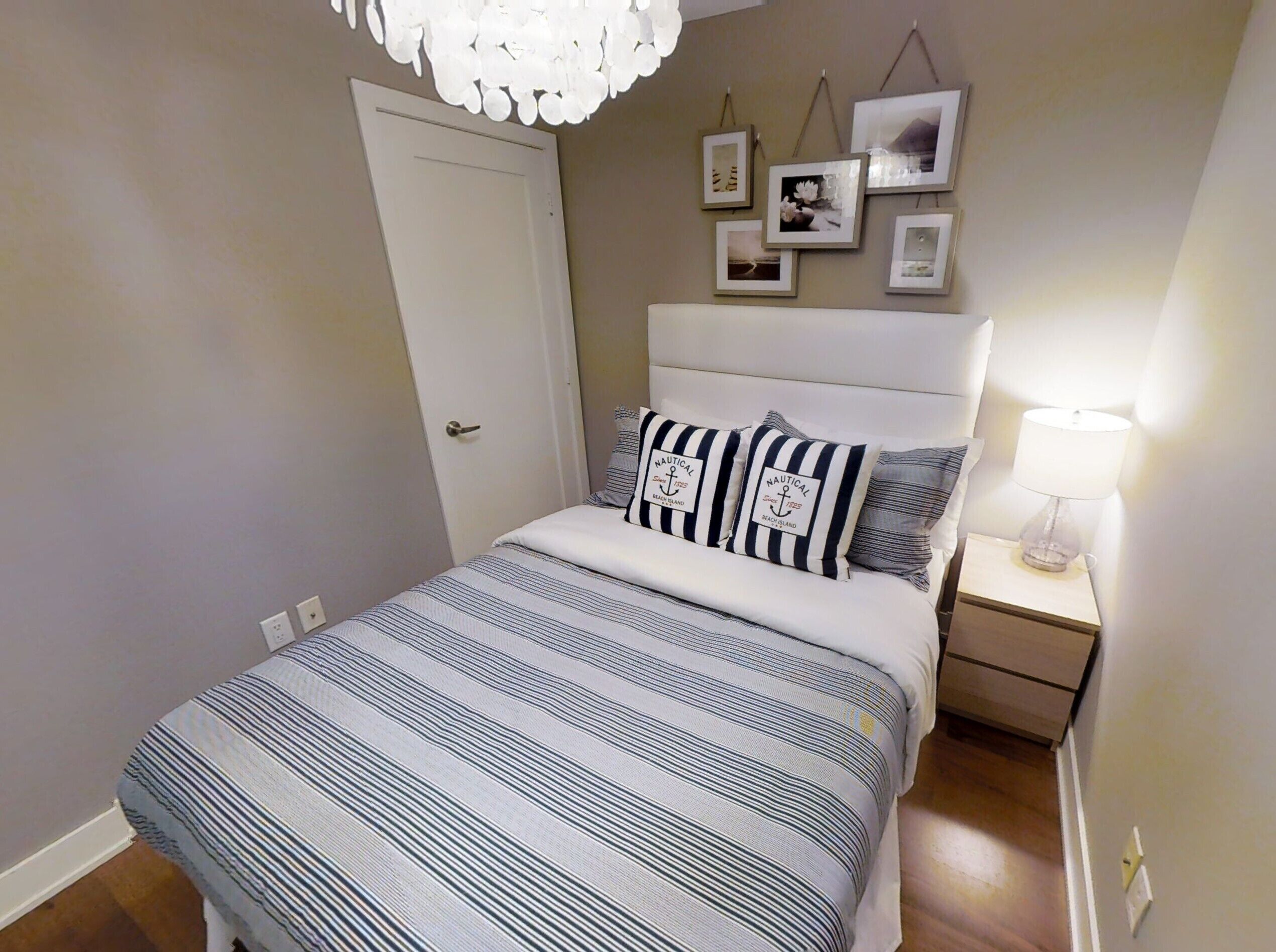 Cama bem arrumada com roupa de cama listrada, mesa de cabeceira com abajur e parede com quadros.