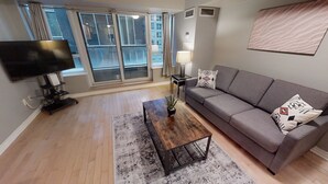 Ein modernes Wohnzimmer mit einem grauen Sofa, einem Holztisch, einem Fernseher und einem großen Fenster mit Vorhängen.