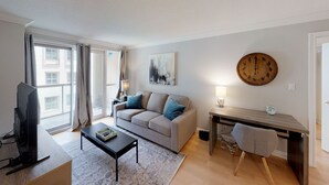 A modern living room with a grey sofa, a coffee table, a TV, a wooden desk, and a large wall clock. - Centrally Located 2-BR Condo in DT Toronto (Toronto)