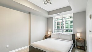 A modern bedroom with a large bed, a window with a city view, and two bedside tables with lamps. - Modern 1-BR Condo DT Toronto (Toronto)