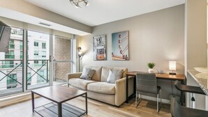 A modern living room with a sofa, coffee table, and a desk with a chair. There are large windows and artwork on the wall. - Modern 1-BR Condo DT Toronto (Toronto)