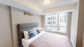 A modern bedroom with a large bed, a headboard, a bedside table with a lamp, and a window with a city view.