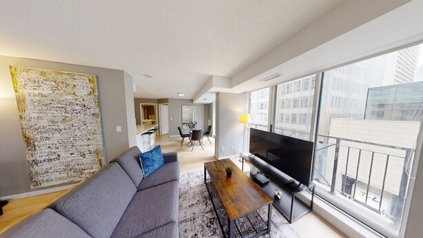 A modern living room with a grey sofa, a coffee table, and a TV. There is a large window with a view of buildings.