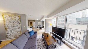 Una sala de estar moderna con un sofá gris, una mesa de centro y un televisor. Hay un ventanal con vistas a edificios.