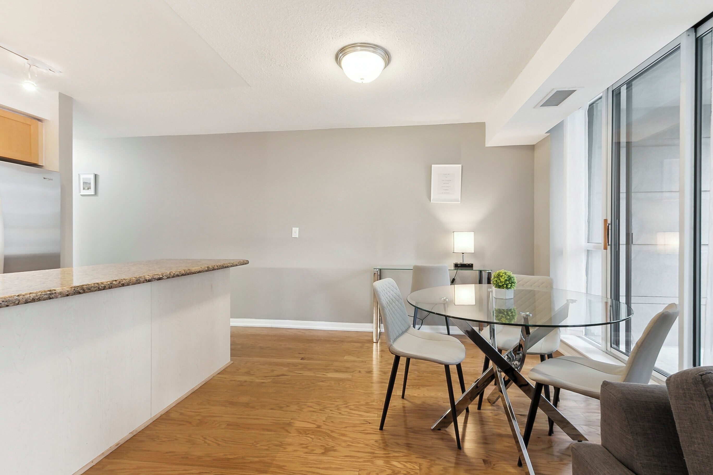 A modern kitchen with a granite countertop, a dining area with a glass table and chairs, and a large window providing natural light.