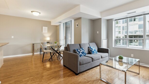 A modern living room with a grey sofa, a glass coffee table, and a dining area with a glass table and chairs. - Modern 1+1-BR Condo DT Toronto (Toronto)