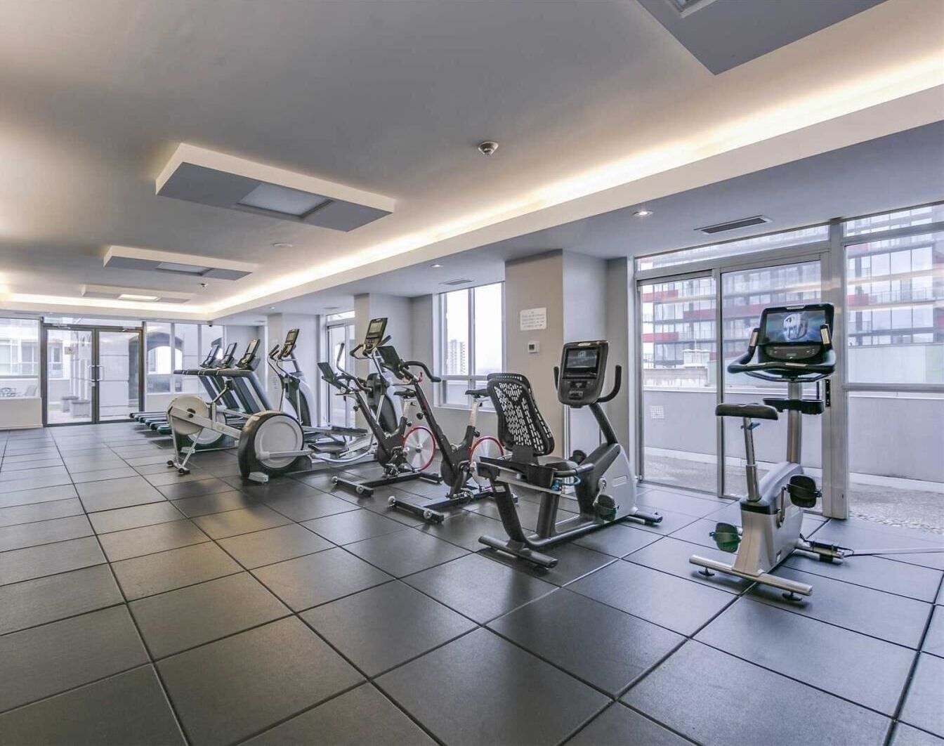 Ein modernes Fitnessstudio mit verschiedenen Cardiogeräten, großen Fenstern und einem sauberen, gefliesten Boden.