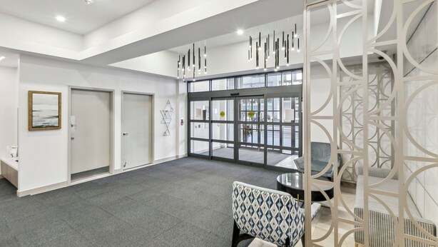 Un hall d’entrée spacieux avec un plafond haut, des luminaires modernes, une porte d’entrée en verre et un coin salon agrémenté de fauteuils à motifs.
