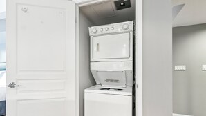 Une petite buanderie avec une machine à laver et un sèche-linge superposés, une porte blanche et un mur de couleur gris clair.