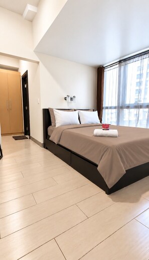 Habitación de hotel moderna con una cama grande, una mesita de noche, una ventana con cortinas y una puerta que conduce a otra habitación.