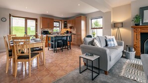 A spacious living area with a grey sofa, wooden dining table, and chairs. A kitchen area is visible with wooden cabinets and a central island. - 2 Bedroom Home in Kenmare With sea Views to Sleep 4 (Kenmare)