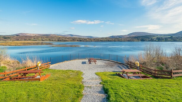 A lakeside view with a wooden deck, picnic tables, and a gravel path leading to a bench. - 2 Bedroom Home in Kenmare With sea Views to Sleep 4 (Kenmare)