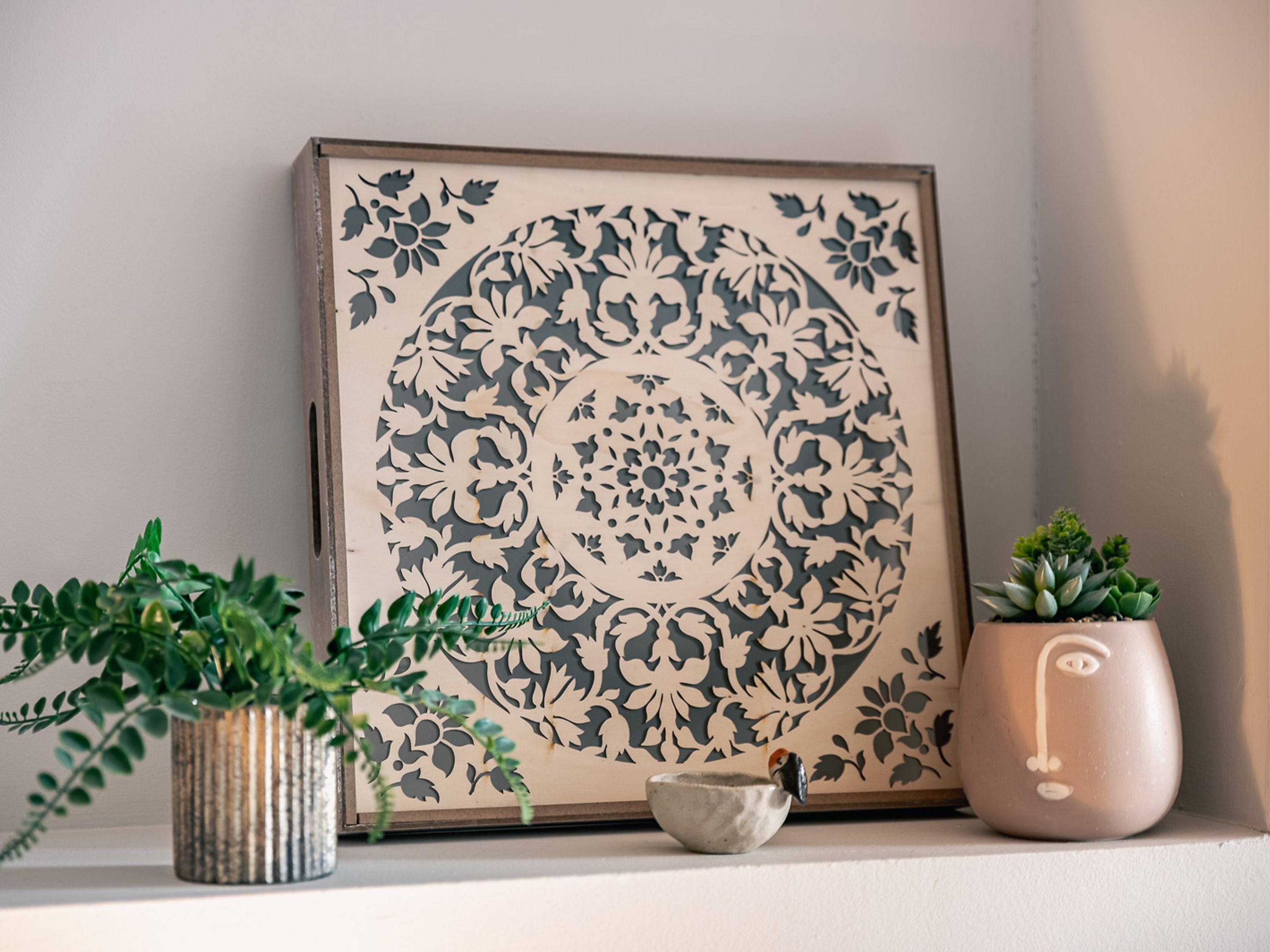 Une œuvre d’art encadrée ornée d’un motif floral, une plante en pot, un pot en céramique avec un visage et un petit bol posé sur une étagère.