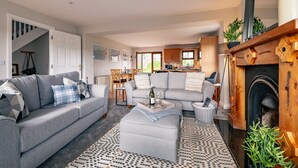 A spacious living room with a grey sofa, ottoman, and a coffee table with a bottle and glasses. There is a fireplace with a mirror above it and a dining area in the background. - 2 Bedroom Home in Kenmare With sea Views to Sleep 4 (Kenmare)