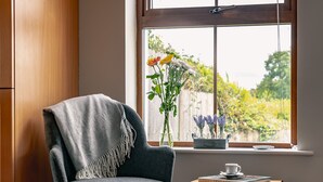 A room with a grey armchair, a small wooden table with a book and a cup, and a window with a view of greenery. - 2 Bedroom Home in Kenmare With sea Views to Sleep 4 (Kenmare)