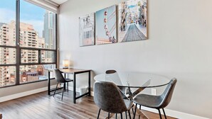 Un espace repas moderne avec une table et des chaises en verre, de grandes fenêtres offrant une vue sur la ville et trois œuvres d’art encadrées accrochées au mur.