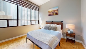 A bedroom with a bed, a nightstand, a lamp, a window with blinds, and a cityscape painting on the wall. - Lovely 1-BR in DT Toronto (Toronto)