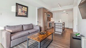 A modern living room with a grey sofa, wooden coffee table, and a kitchen area with a fridge, microwave, and oven. - Lovely 1-BR in DT Toronto (Toronto)