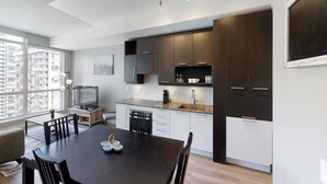 Et moderne køkken med mørke skabe, et spiseområde med bord og stole, samt et stort vindue med udsigt over bybilledet.