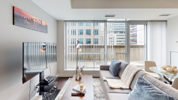 A modern living room with a flat-screen TV, a sofa, a coffee table, and a large window with blinds. - Modern Central 1-BR Condo in DT Toronto (Toronto)