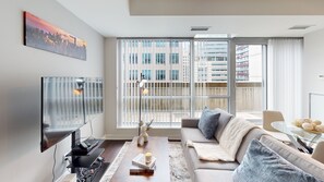 Een moderne woonkamer met een flatscreen televisie, een bank, een salontafel en een groot raam met zonwering.