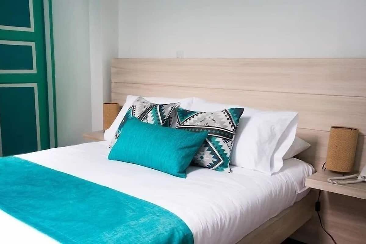 Una cama bien hecha con una manta color turquesa, ropa de cama blanca y almohadas estampadas. Una mesita de noche de madera con un teléfono y una lámpara está al lado de la cama.