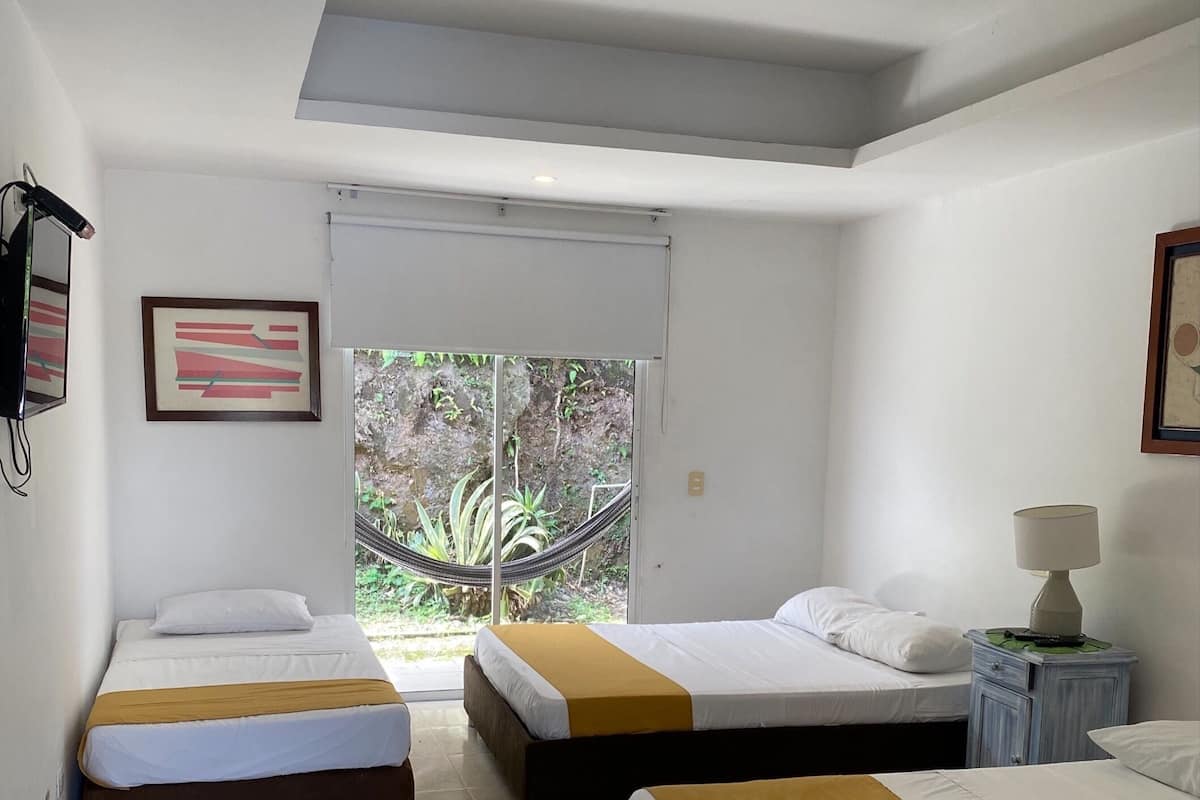 Habitación de hotel con dos camas, una mesita con una lámpara y una ventana con vistas a zonas verdes.