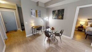 A modern dining area with a round table, four chairs, and a wooden side table with a lamp. There are two paintings on the walls and a bed with a nightstand in the adjacent room. - Modern Central 1+1-BR Condo in DT Toronto (Toronto)