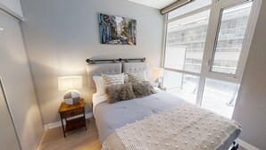 A bedroom with a large bed, a nightstand with a lamp, a painting on the wall, and a window with a view of buildings. - Modern Central 1+1-BR Condo in DT Toronto (Toronto)