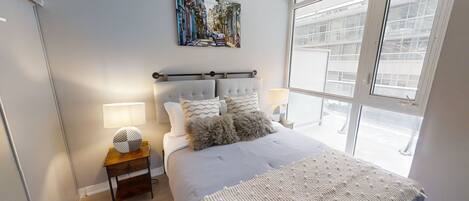 Un dormitorio con una cama grande, una mesita de noche con lámpara, un cuadro en la pared y una ventana con vistas a edificios.