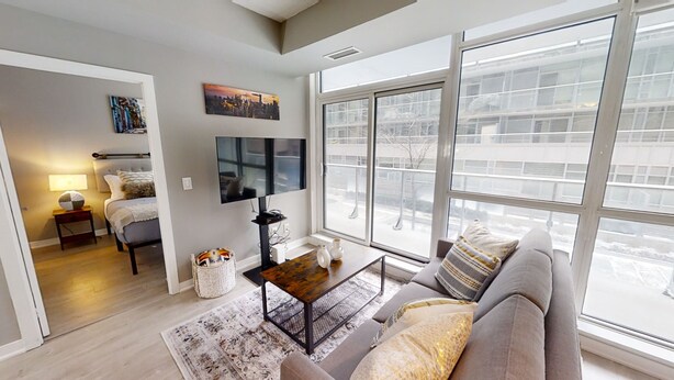 A modern living room with a large window, a flat-screen TV, a sofa, and a coffee table. - Modern Central 1+1-BR Condo in DT Toronto (Toronto)