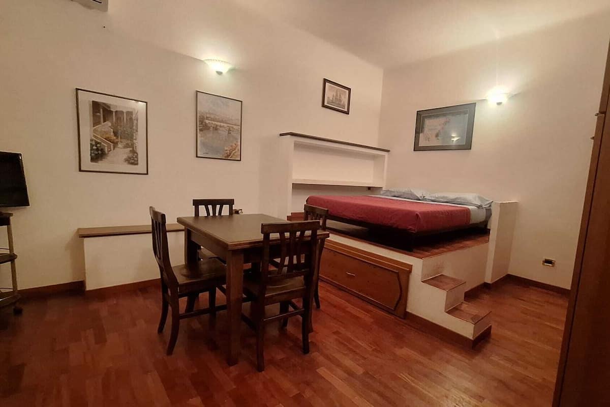 Una habitación pequeña con una mesa de comedor de madera y sillas, una cama litera con colchón rojo y varias fotos enmarcadas en las paredes.