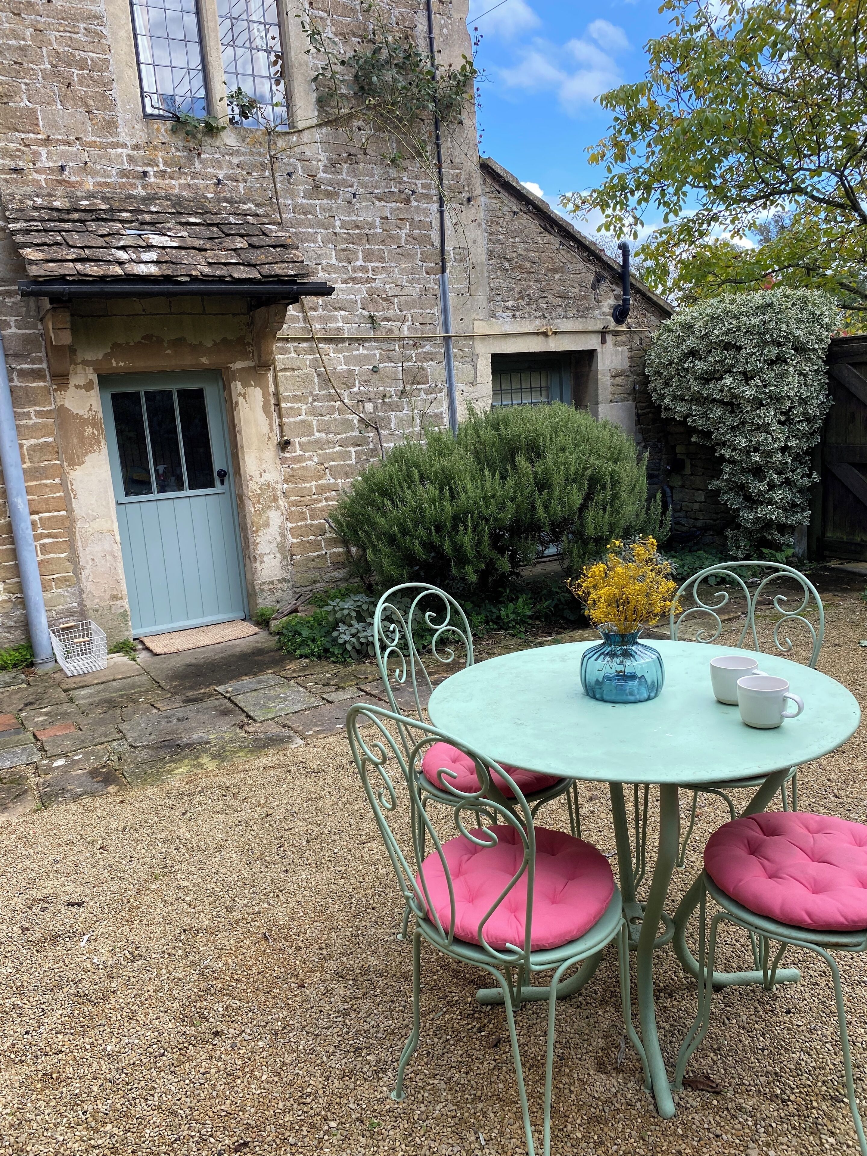 Un bâtiment rustique en pierre, doté d’une porte bleue, un petit jardin avec une table ronde et des chaises en métal, et un arbre aux fleurs jaunes.