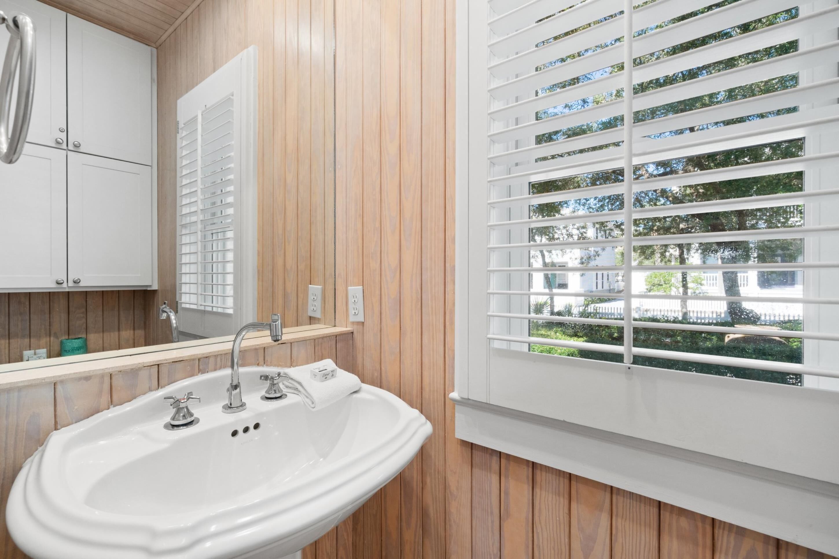 Un baño con un lavamanos blanco, paneles de madera en las paredes, un espejo y una ventana con persianas.