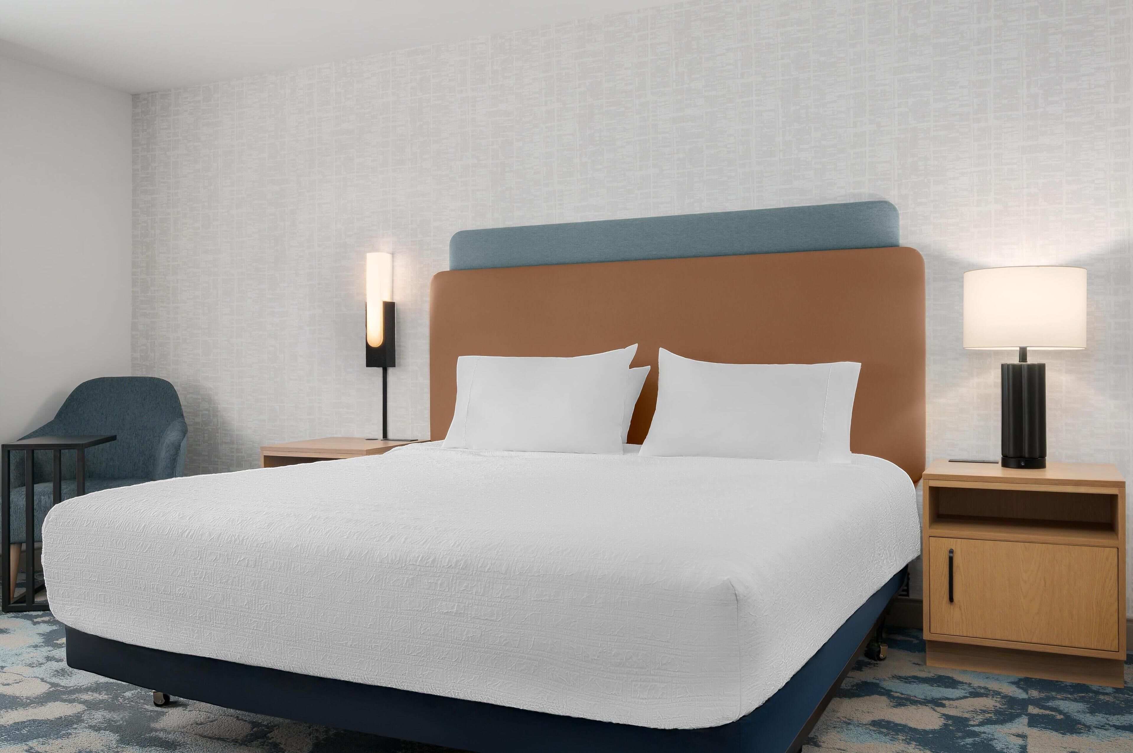 Una habitación de hotel con una cama grande, dos mesitas de noche, una silla y una lámpara de pared.
