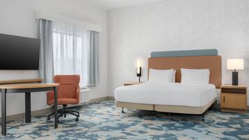 A hotel room with a bed, a desk, a chair, a TV, and a nightstand with a lamp.