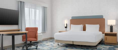 Habitación de hotel con cama, escritorio, silla, televisor y mesita de noche con lámpara.