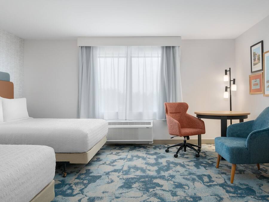 A hotel room with two beds, a standing heater, a blue patterned carpet, and a small round table with a chair.